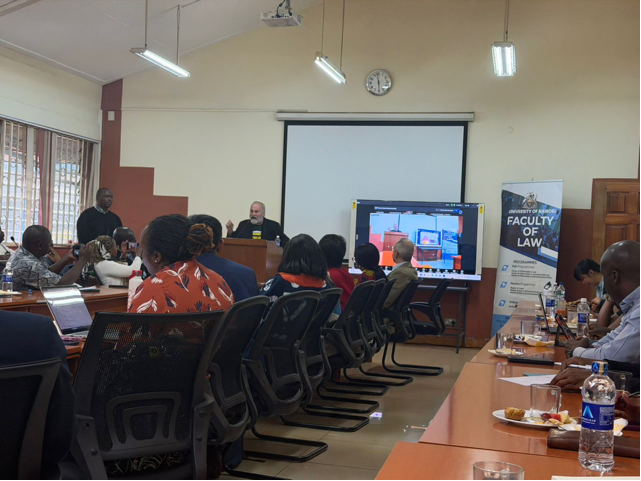 A group of people sit in a conference room watching Robert Percival speak at a podium.